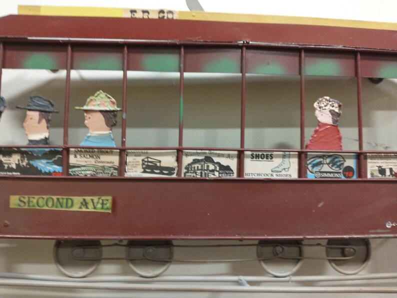 May include: A red and brown metal model of a streetcar with a green and white interior. The streetcar has a sign that reads "Second Ave" and has several small advertisements on the side. The streetcar has four wheels and is on a track.