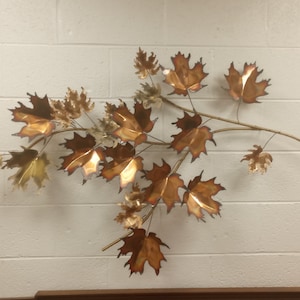 Puede incluir: Una escultura de pared que presenta una rama con m&uacute;ltiples hojas de arce de color cobre. Las hojas est&aacute;n dispuestas en un patr&oacute;n en cascada, creando una sensaci&oacute;n de movimiento y belleza natural.