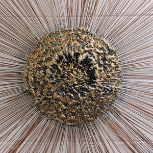 Puede incluir: Una escultura de pared de metal dorado con un centro circular texturizado y finos alambres met&aacute;licos que irradian.