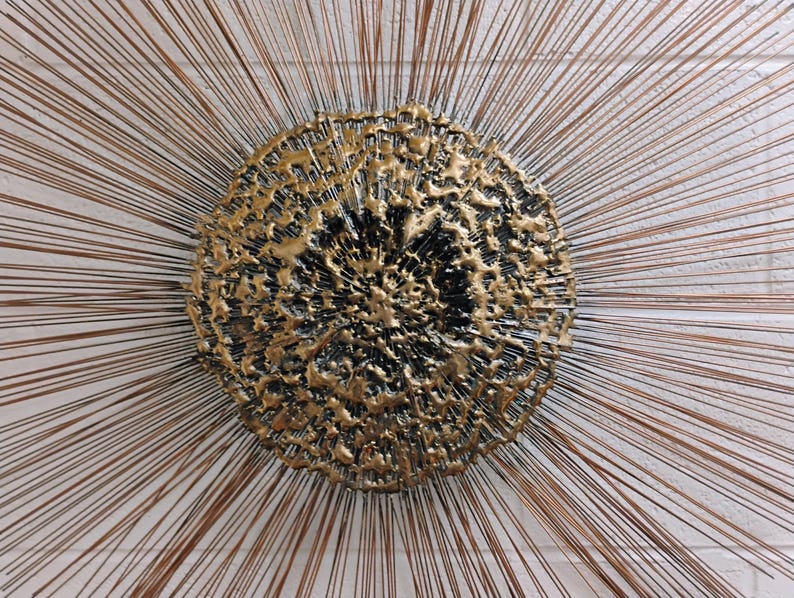 Puede incluir: Una escultura de pared de metal dorado con un centro circular texturizado y finos alambres met&aacute;licos que irradian.