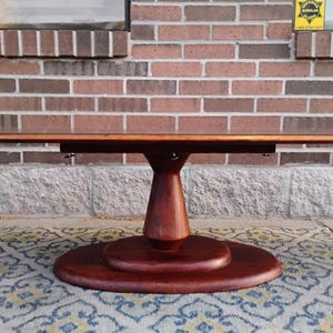 May include: A round, dark-stained wooden table with a pedestal base. The tabletop is rectangular and extends outward. The table is set on a patterned rug, with a brick wall in the background. The table is approximately 60 inches long.