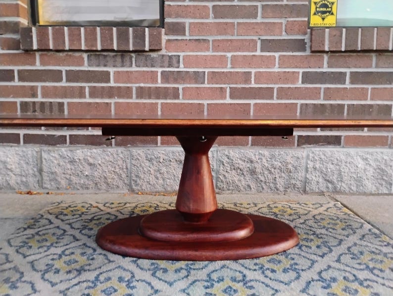 May include: A round, dark-stained wooden table with a pedestal base. The tabletop is rectangular and extends outward. The table is set on a patterned rug, with a brick wall in the background. The table is approximately 60 inches long.