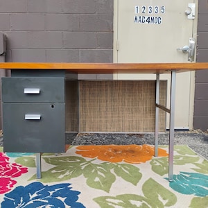 May include: A desk featuring a rectangular wood-tone top, a dark grey metal cabinet with two drawers and silver-toned horizontal handles, and a silver-toned metal leg frame. The desk includes a textured, light brown privacy panel at the back.