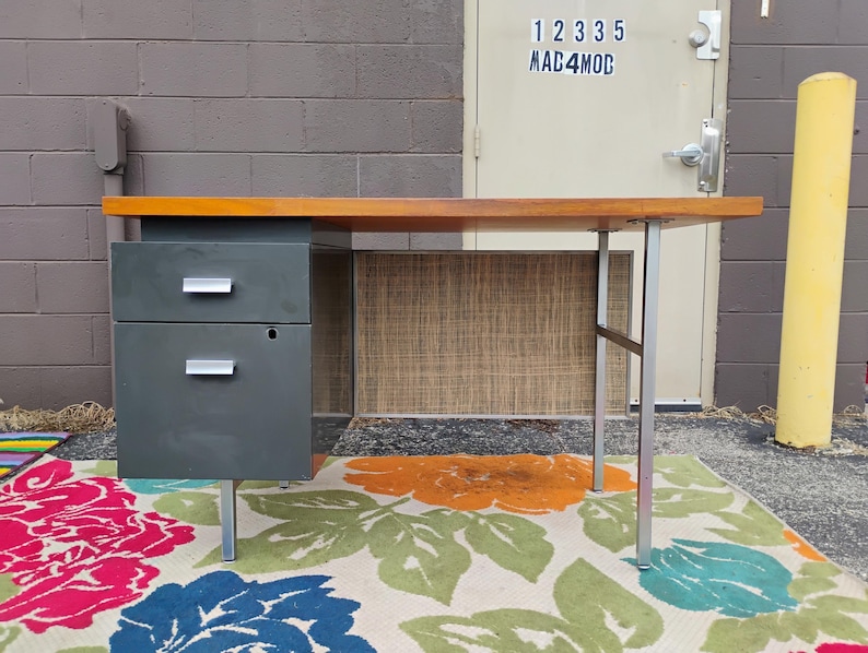 May include: A desk featuring a rectangular wood-tone top, a dark grey metal cabinet with two drawers and silver-toned horizontal handles, and a silver-toned metal leg frame. The desk includes a textured, light brown privacy panel at the back.
