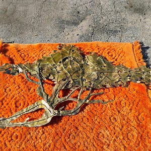 May include: A large, gold-coloured metal wall sculpture depicting a stylised tree with intricate leaf details. The sculpture is lying on an orange rug.