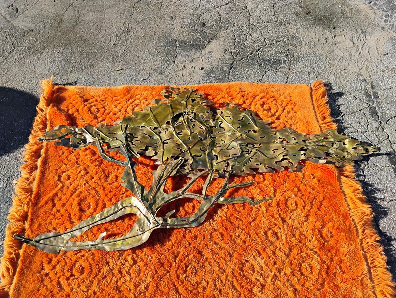 May include: A large, gold-coloured metal wall sculpture depicting a stylised tree with intricate leaf details. The sculpture is lying on an orange rug.