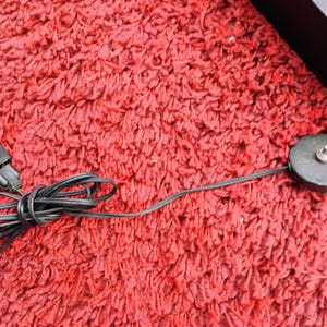 K&ouml;nnte beinhalten: Ein schwarzes Elektrokabel mit einem gr&uuml;nspitzigen Stecker und einem kreisf&ouml;rmigen Ein/Aus-Schalter. Das Kabel ist aufgerollt und liegt auf einer roten, strukturierten Oberfl&auml;che. Der Schalter ist dunkelgrau mit einem kleinen, silbernen Knopf.