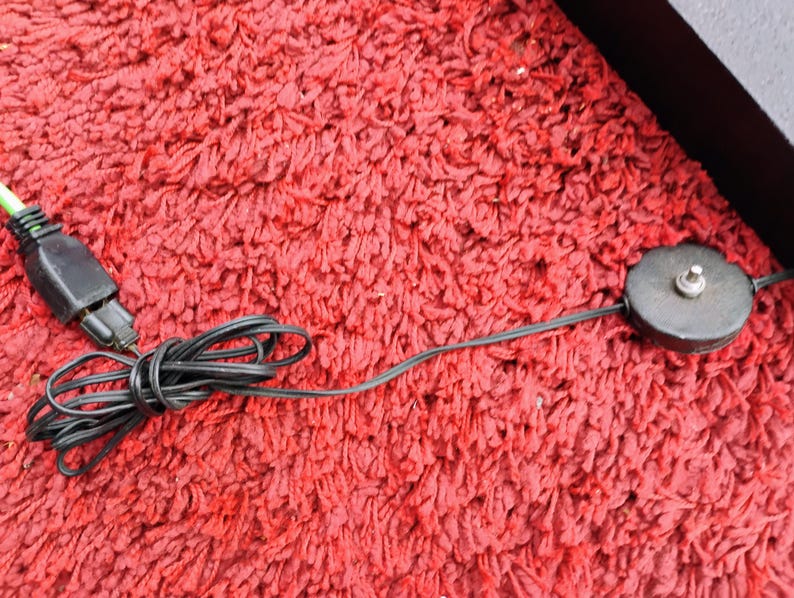 K&ouml;nnte beinhalten: Ein schwarzes Elektrokabel mit einem gr&uuml;nspitzigen Stecker und einem kreisf&ouml;rmigen Ein/Aus-Schalter. Das Kabel ist aufgerollt und liegt auf einer roten, strukturierten Oberfl&auml;che. Der Schalter ist dunkelgrau mit einem kleinen, silbernen Knopf.
