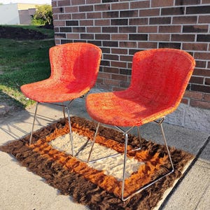 May include: Two mid-century modern chairs with bright orange textured upholstery. The chairs have a curved back and seat, supported by slender, silver-colored metal legs. They sit on a brown, orange, and cream-colored rug, with a brick wall in the background.