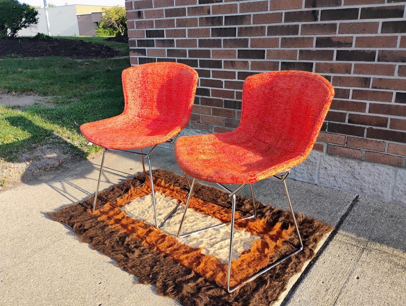 May include: Two mid-century modern chairs with bright orange textured upholstery. The chairs have a curved back and seat, supported by slender, silver-colored metal legs. They sit on a brown, orange, and cream-colored rug, with a brick wall in the background.
