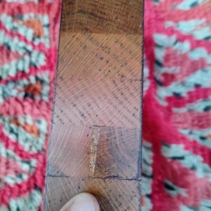May include: Close-up of a section of a wooden object, possibly a table leg or a similar furniture component. The wood is a warm brown color, with visible grain patterns and a slightly rough texture. The object is composed of several rectangular blocks of wood.