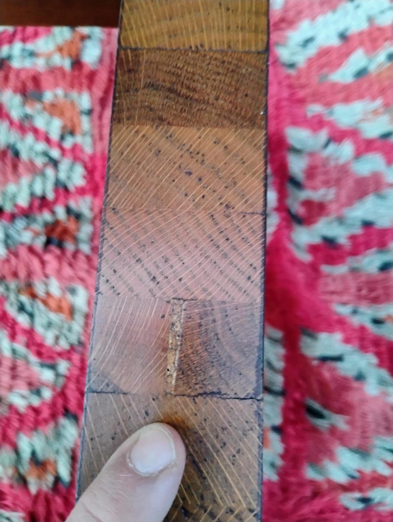 May include: Close-up of a section of a wooden object, possibly a table leg or a similar furniture component. The wood is a warm brown color, with visible grain patterns and a slightly rough texture. The object is composed of several rectangular blocks of wood.