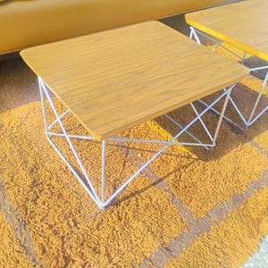 May include: Two square side tables with wood grain laminate tops and white metal wire bases. The tables feature a geometric frame design with thin metal rods forming a crisscross pattern on each side, supporting the flat, square wooden surfaces.