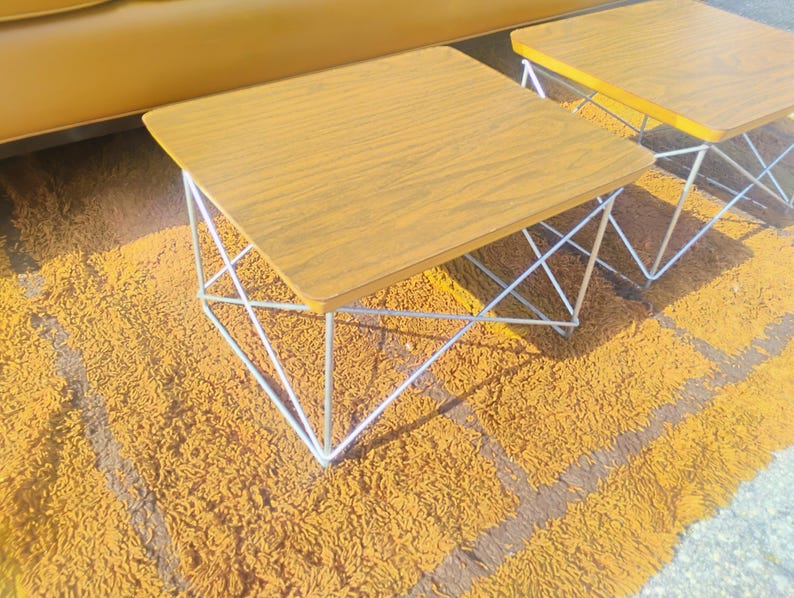 May include: Two square side tables with wood grain laminate tops and white metal wire bases. The tables feature a geometric frame design with thin metal rods forming a crisscross pattern on each side, supporting the flat, square wooden surfaces.
