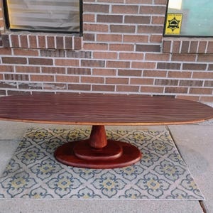 May include: Oval-shaped wooden coffee table with a dark brown finish. The table has a pedestal base and sits on a patterned rug. The table's dimensions are approximately 60 inches long and 24 inches wide.