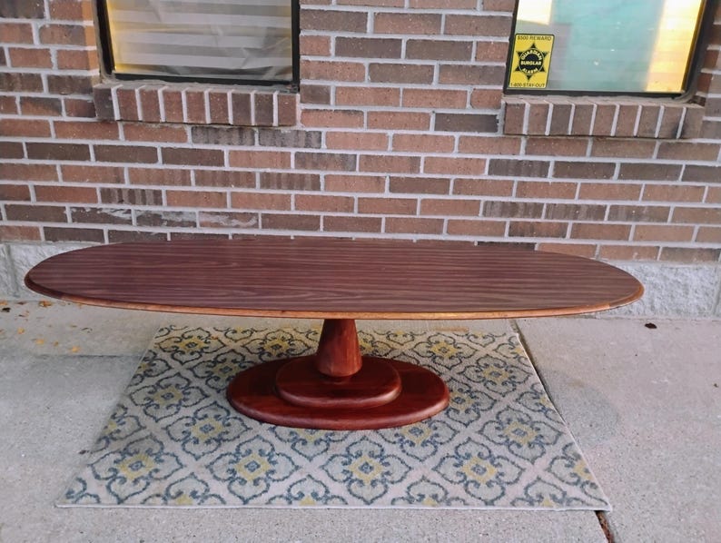 May include: Oval-shaped wooden coffee table with a dark brown finish. The table has a pedestal base and sits on a patterned rug. The table's dimensions are approximately 60 inches long and 24 inches wide.