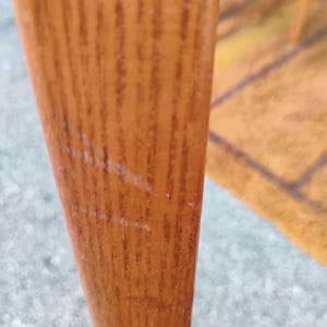 May include: A vertical wooden furniture leg with a light brown finish and a visible natural grain pattern. The surface shows light scratches and wear marks along the length of the wood, highlighting the texture and material of the furniture component.