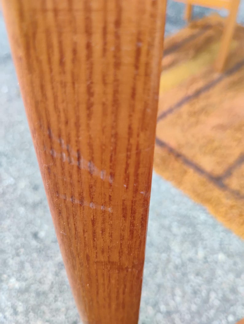 May include: A vertical wooden furniture leg with a light brown finish and a visible natural grain pattern. The surface shows light scratches and wear marks along the length of the wood, highlighting the texture and material of the furniture component.