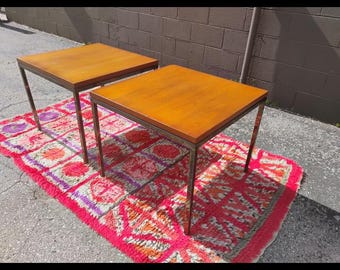 Könnte beinhalten: Zwei quadratische Beistelltische aus Holz mit Metallbeinen. Die Tische haben eine warme Holzoberfläche und stehen auf einem gemusterten Teppich mit rosa, roten und violetten Farbtönen. Eine stilvolle Ergänzung für jeden Wohnraum.