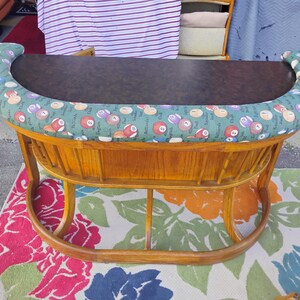 May include: A vintage, wooden pool table with a dark brown playing surface. The table's padded edge is covered in green fabric with a repeating pattern of billiard balls. The table has a curved base and legs.