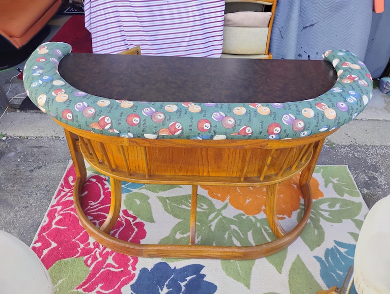 May include: A vintage, wooden pool table with a dark brown playing surface. The table's padded edge is covered in green fabric with a repeating pattern of billiard balls. The table has a curved base and legs.