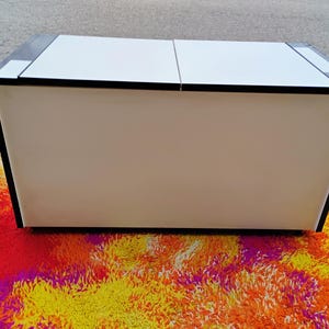 May include: A white rectangular storage cabinet with black trim. The cabinet has two doors and sits on a colorful rug.