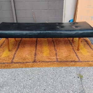 May include: A rectangular black bench featuring a tufted seat with six buttons and four tapered wooden legs. The bench has a smooth, dark surface and a low-profile design, supported by four light-colored wooden legs positioned at each corner.