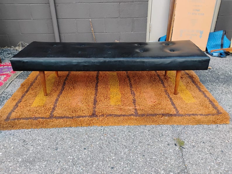 May include: A rectangular black bench featuring a tufted seat with six buttons and four tapered wooden legs. The bench has a smooth, dark surface and a low-profile design, supported by four light-colored wooden legs positioned at each corner.