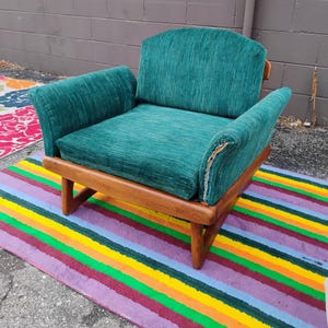 May include: A low-profile armchair featuring a wooden frame with a natural finish and a textured, deep green fabric upholstery. The chair has a wide seat, a curved backrest, and integrated armrests, with visible wear on the right armrest fabric.