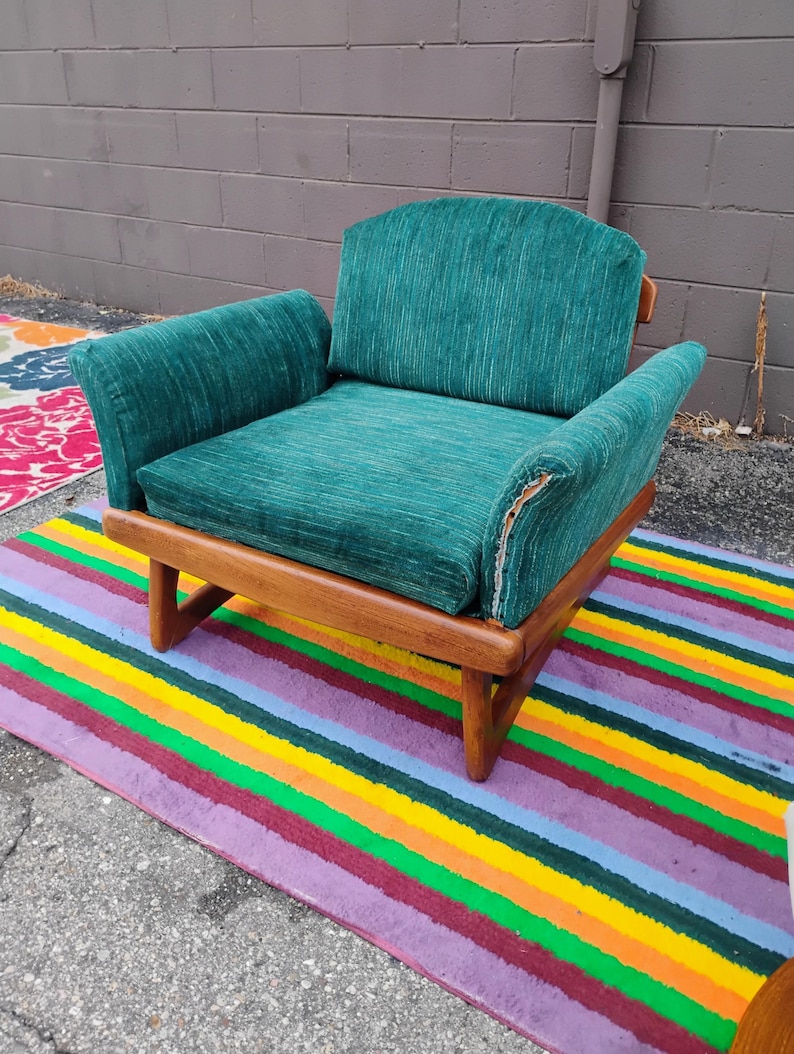 May include: A low-profile armchair featuring a wooden frame with a natural finish and a textured, deep green fabric upholstery. The chair has a wide seat, a curved backrest, and integrated armrests, with visible wear on the right armrest fabric.