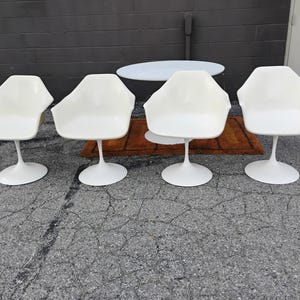 May include: A set of four white molded plastic armchairs, each featuring a single central pedestal base. The chairs have a smooth, curved shell design with integrated armrests and a wide, circular base for stability on the ground.