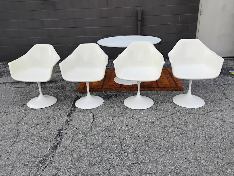 May include: A set of four white molded plastic armchairs, each featuring a single central pedestal base. The chairs have a smooth, curved shell design with integrated armrests and a wide, circular base for stability on the ground.