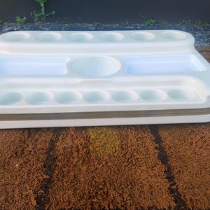 May include: A white plastic organizer tray featuring a tiered design with multiple circular indentations along the top and bottom edges, two rectangular recessed compartments, and a central circular basin, accented by a horizontal metallic band.