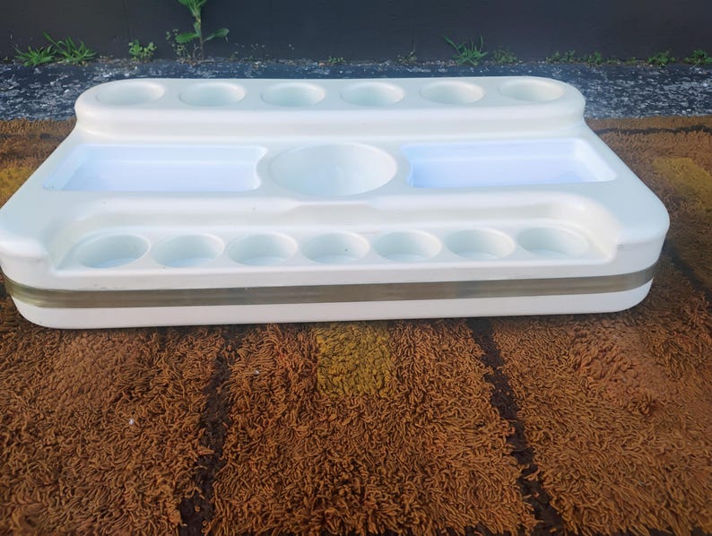 May include: A white plastic organizer tray featuring a tiered design with multiple circular indentations along the top and bottom edges, two rectangular recessed compartments, and a central circular basin, accented by a horizontal metallic band.