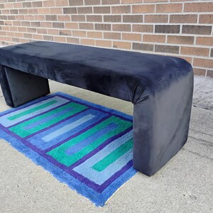 May include: A navy blue upholstered bench with a rectangular shape and rounded corners. The bench is placed over a rectangular rug with blue, green, and purple stripes. The bench is approximately 60 inches long.