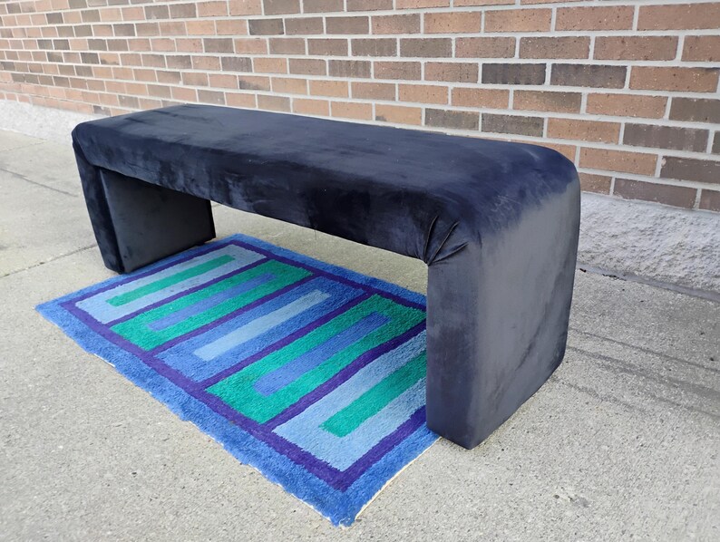 May include: A navy blue upholstered bench with a rectangular shape and rounded corners. The bench is placed over a rectangular rug with blue, green, and purple stripes. The bench is approximately 60 inches long.