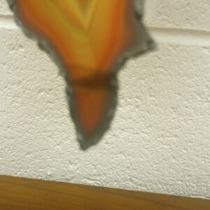 Puede incluir: Un primer plano de un adorno de metal en forma de llama con una superficie texturizada. El adorno est&aacute; pintado en tonos de naranja y amarillo, con un naranja m&aacute;s oscuro en la punta. El adorno est&aacute; colgado de una pared blanca.