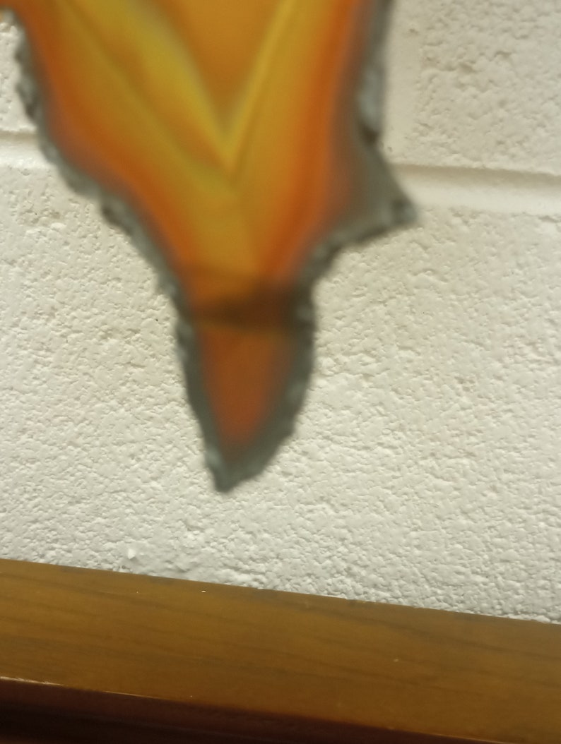 Puede incluir: Un primer plano de un adorno de metal en forma de llama con una superficie texturizada. El adorno est&aacute; pintado en tonos de naranja y amarillo, con un naranja m&aacute;s oscuro en la punta. El adorno est&aacute; colgado de una pared blanca.