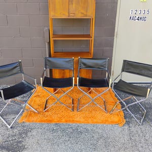 May include: Four folding chairs with black leather seats and backrests, featuring polished chrome metal frames. The chairs are arranged in a row on an orange textured rug in front of a tall wooden shelving unit with two cabinet doors at the top.