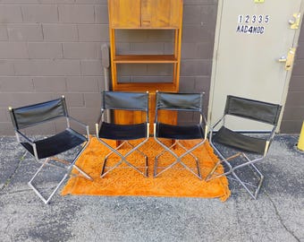 May include: Four folding chairs with black leather seats and backrests, featuring polished chrome metal frames. The chairs are arranged in a row on an orange textured rug in front of a tall wooden shelving unit with two cabinet doors at the top.