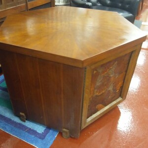 May include: A mid-century modern hexagonal side table made of wood. The table has a rich brown finish with a patterned top and a decorative panel on one side. The table is supported by short, rectangular legs.