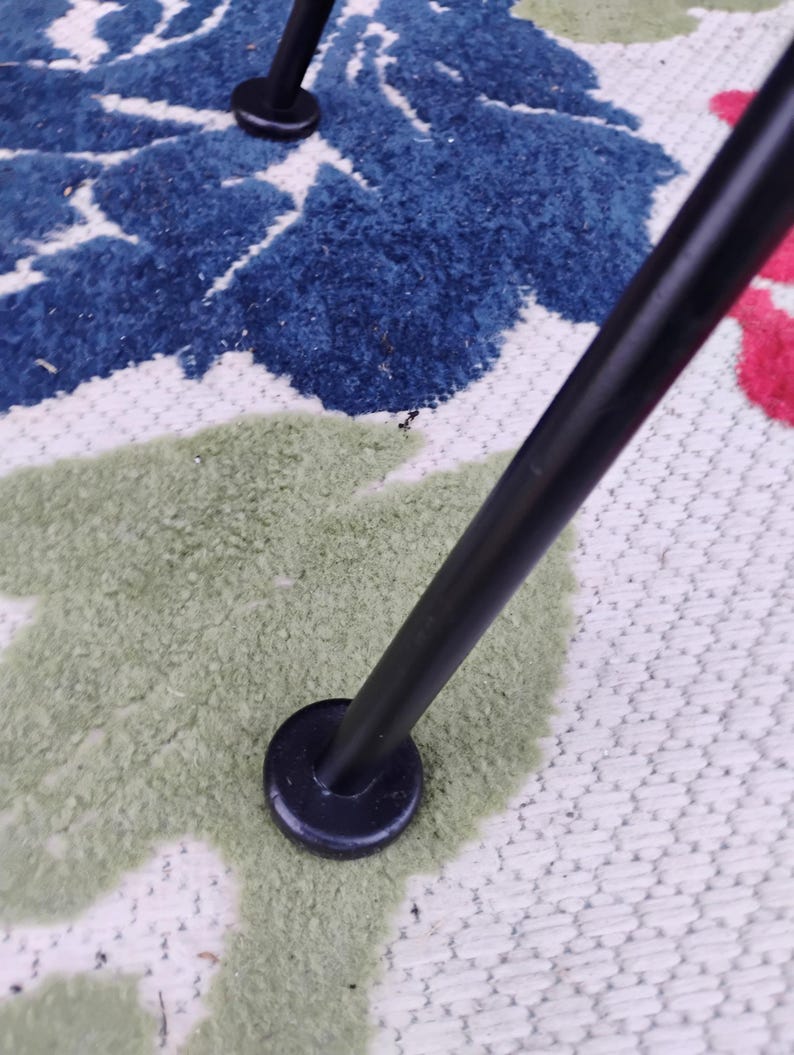 May include: Close-up of a black metal chair leg with a round black foot pad. The chair leg is resting on a white and green patterned rug.