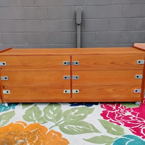 May include: A rectangular wooden dresser with a warm brown wood grain finish, featuring six drawers arranged in two vertical columns. Each drawer is equipped with a flat, silver-toned metal pull handle on the left and right sides.