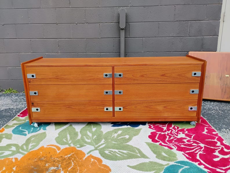 May include: A rectangular wooden dresser with a warm brown wood grain finish, featuring six drawers arranged in two vertical columns. Each drawer is equipped with a flat, silver-toned metal pull handle on the left and right sides.