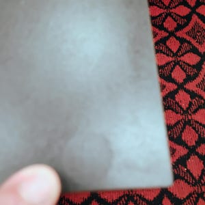 May include: A rectangular, dark gray, matte-finished object with a smooth, solid surface. The item features a subtle, circular discoloration or mark on its lower portion, presenting a uniform, flat appearance across the main body of the piece.