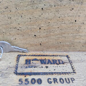 May include: A set of silver keys on a ring, resting on a wooden surface. The keys are next to a rectangular, dark-colored stamp that reads "HOWARD" and "5500 GROUP". The wood has a worn, textured appearance.