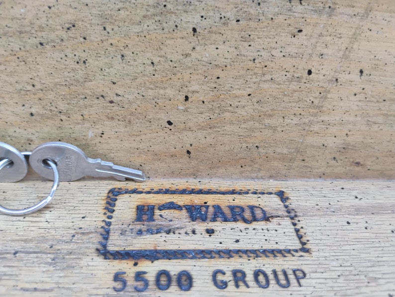 May include: A set of silver keys on a ring, resting on a wooden surface. The keys are next to a rectangular, dark-colored stamp that reads "HOWARD" and "5500 GROUP". The wood has a worn, textured appearance.