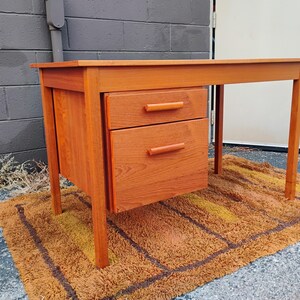 Op de afbeelding: Een rechthoekig houten bureau met een warme oranjebruine afwerking, voorzien van een vlak blad, vier rechte poten en een zijblok met een kleinere bovenste lade en een grotere onderste lade, beide voorzien van een horizontale houten handgreep.