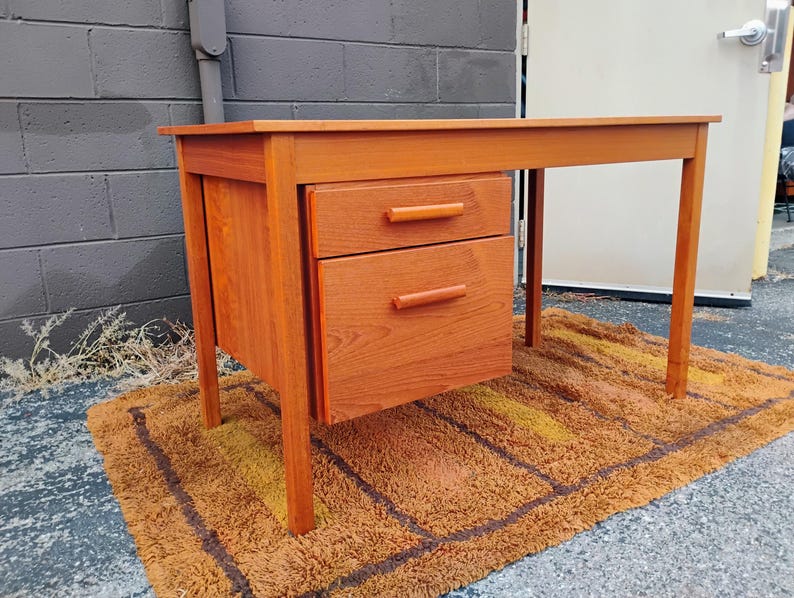 Op de afbeelding: Een rechthoekig houten bureau met een warme oranjebruine afwerking, voorzien van een vlak blad, vier rechte poten en een zijblok met een kleinere bovenste lade en een grotere onderste lade, beide voorzien van een horizontale houten handgreep.