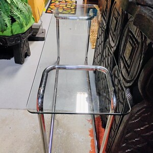 Peut inclure: Table d'appoint vintage en chrome et verre, avec deux &eacute;tag&egrave;res rectangulaires en verre. La table a un design &eacute;pur&eacute; et minimaliste avec des pieds et des supports chrom&eacute;s incurv&eacute;s. Les &eacute;tag&egrave;res en verre sont transparentes, offrant une esth&eacute;tique moderne.